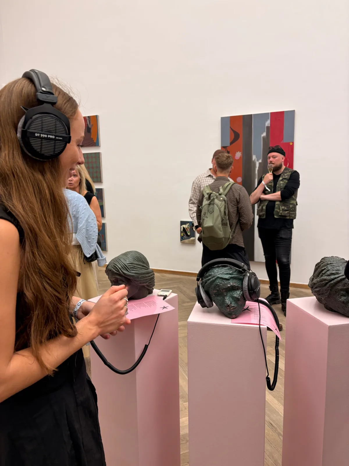 Visitors wearing headphones beside sculptures and paintings in an exhibition.